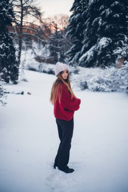 Kadın kışın ağaçlarla karlı bir arazide duruyor, sıcak giysiler ve gün boyunca örülmüş bir şapka giyiyor.