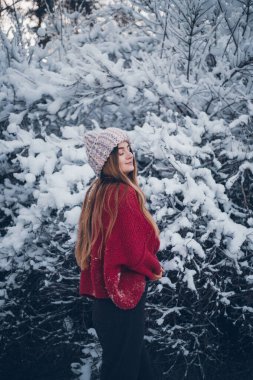Genç bir kadın, ormanda serin bir günde, kırmızı bir kazak ve kış şapkası giymiş, karla kaplı ağaçların yanında duruyor.