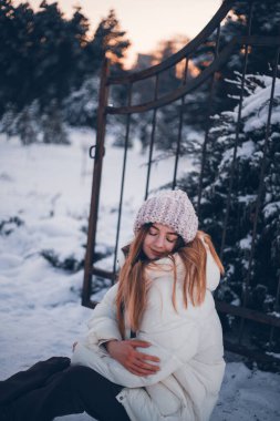 Genç bir kadın, kışın gün batımında ağaçların yanında, karla çevrili bir kapıda oturur.