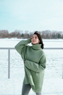 Yeşil kazaklı kadın, kışın öğleden sonra donmuş gölün yanında, güpegündüz, karlı sahilde poz veriyor.