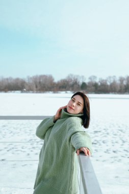 Yeşil kazaklı kadın, kışın öğleden sonra donmuş gölün yanında, güpegündüz, karlı sahilde poz veriyor.