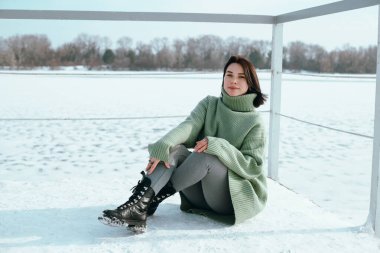 Kadın kışın donmuş gölün yanındaki karla kaplı zeminde yeşil kazak ve botlarıyla oturuyor.