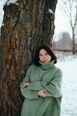 Kadın kış boyunca karlı bir parkta bir ağacın yanında durur. Arka planda ağaçlar vardır. Gün ortasında.
