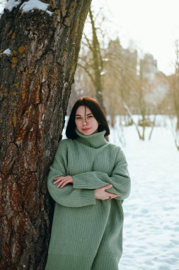 Kadın kış boyunca karlı bir parkta bir ağacın yanında durur. Arka planda ağaçlar vardır. Gün ortasında.