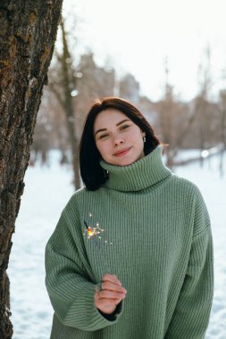 Kışın ayakta duran kadın bir parkta güneşli bir günde elinde bir maytapla gülümsüyor.