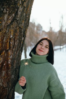 Kışın ayakta duran kadın bir parkta güneşli bir günde elinde bir maytapla gülümsüyor.