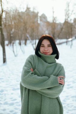 Kadın, kış öğleden sonrası boyunca arka planda ağaçlar ve binalar olan yeşil bir kazak giyerek karlı parkta duruyor.