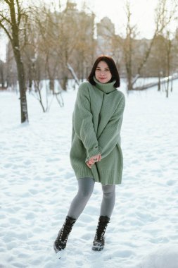 Kadın, kış öğleden sonrası boyunca arka planda ağaçlar ve binalar olan yeşil bir kazak giyerek karlı parkta duruyor.