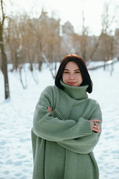 Kadın, kış öğleden sonrası boyunca arka planda ağaçlar ve binalar olan yeşil bir kazak giyerek karlı parkta duruyor.