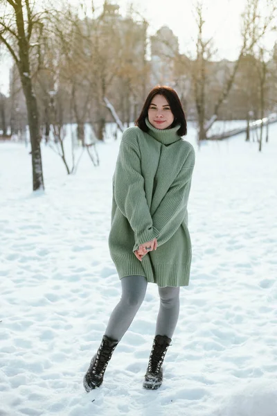 Kadın, kış öğleden sonrası boyunca arka planda ağaçlar ve binalar olan yeşil bir kazak giyerek karlı parkta duruyor.