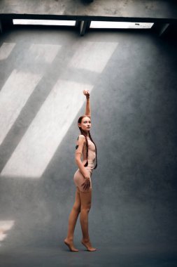 Bir kadın çıplak bir odada tek kolu havada dururken gündüz vakti tavan pencerelerinden ışık akıyor.