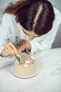 Şekerci, gün ışığında mutfak ortamında kremayla pasta süslüyor.