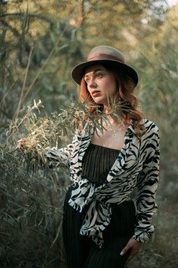 Kadın, güz mevsiminde bir dala dokunurken şapkası ve hayvan desenli kıyafetiyle yeşil ormanda duruyor.