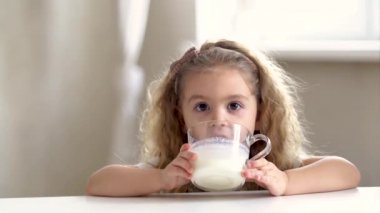 Sevimli sarışın küçük kız mutfakta oturuyor, elinde bardakla süt içiyor ya da yoğurt yiyor sabah içkisinin tadını çıkarıyor kameraya gülümsüyor, çocuk büyütme kavramına bakıyor, sağlıklı yaşam tarzı, sağlık hizmetleri...