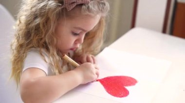 Küçük sarışın kız masada oturuyor, kırmızı bir sevgililer günü kalbi çiziyor. DIY Sevgililer Günü kartı.