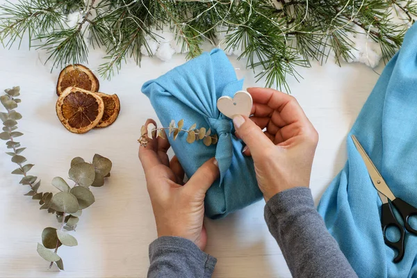 Sevgi dolu Noel hediyeleriyle dolu kadın elleri. El yapımı hediye paketi Japon furoshiki tarzında mavi kumaşa sarılmış. Dekore edilmiş ev masası, üst görünüm.