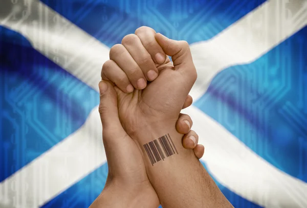 Barcode ID number on wrist of dark skinned person and national flag on background - Scotland — Stock Photo, Image