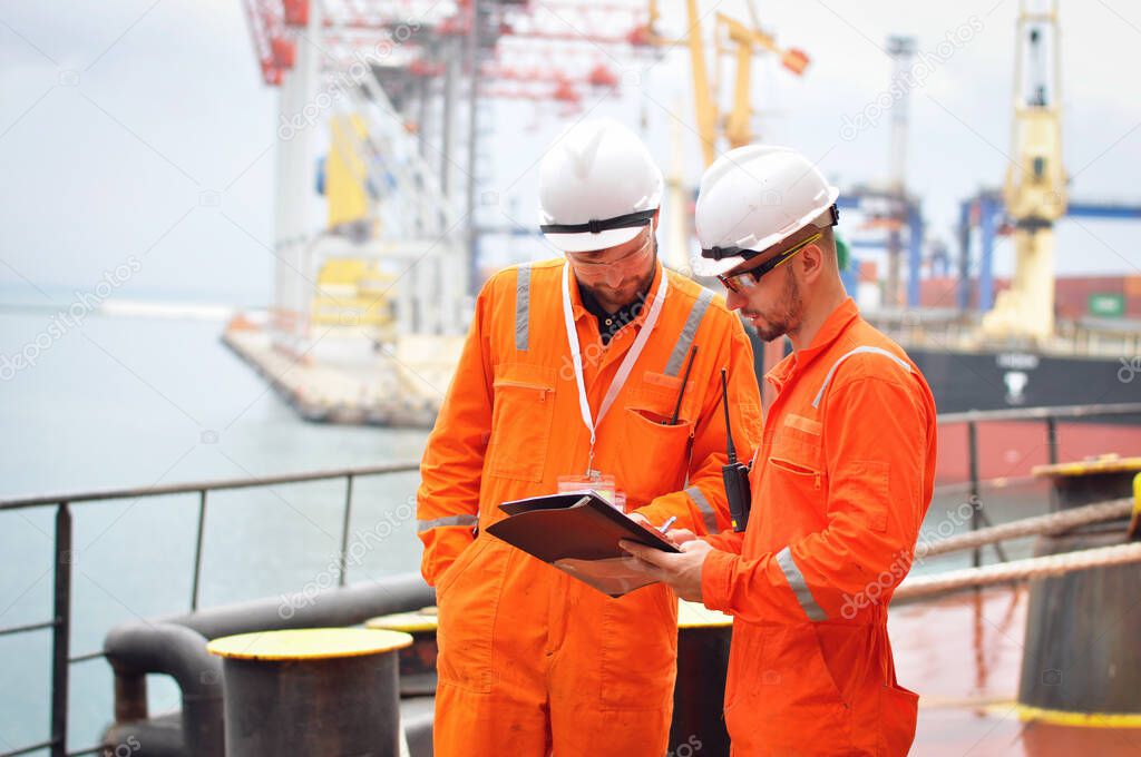 Two sailors in orange overalls work on the ship with documents.