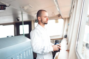 Denizci seyir subayı, kaptanın üçüncü asistanı kaptanlar köprüsünde beyaz tulumlu dürbünle bakıyor..