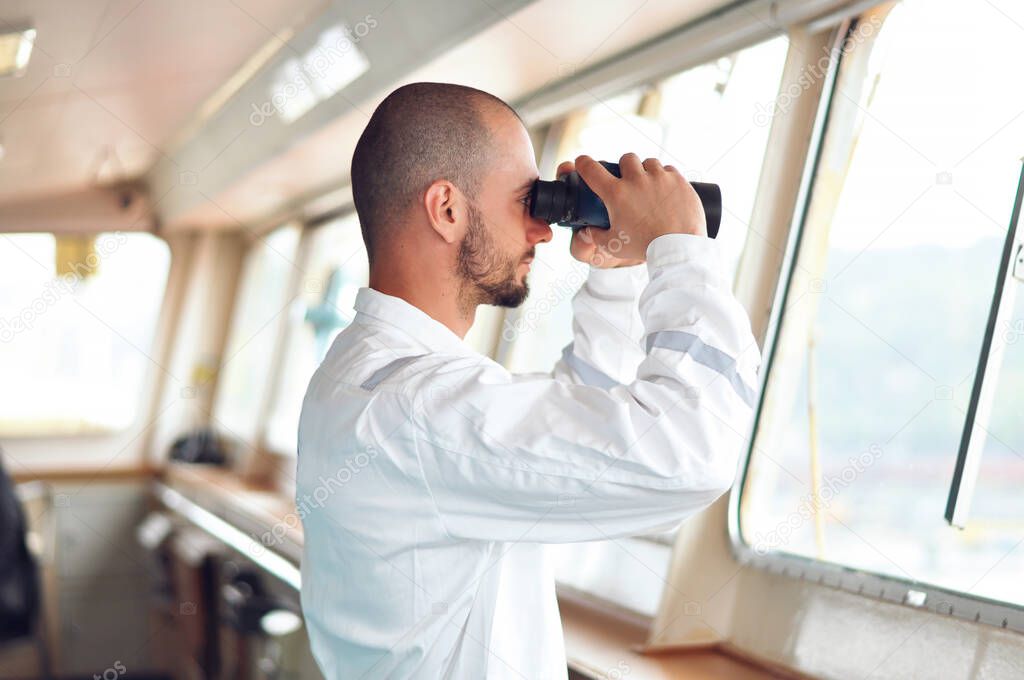 Sailor navigator, the third assistant to the captain looks through binoculars in a white overalls on the captains bridge.