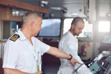 Köprüdeki beyaz gömlekli ve omuz bantlı bir kargo gemisinin kaptanı seyir subayına talimat verir.