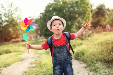Kot pantolonlu, beyaz şapkalı, yaz parkında rüzgar çiçeği oyuncağıyla dolaşan ve dilini gösteren tatlı bir erkek bebek..
