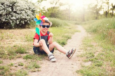 Kot pantolon, güneş gözlüğü ve beyaz şapkalı şirin bir erkek bebek yaz parkında bir patikada oturuyor rüzgar çiçeği oyuncağı ve gülümsüyor..