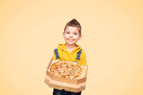 黄色のセーターとデニムでかわいい赤ちゃんの男の子は笑顔をオーバーオールし バックグラウンドで隔離されたピザとオープンボックスを保持しています — ストック写真