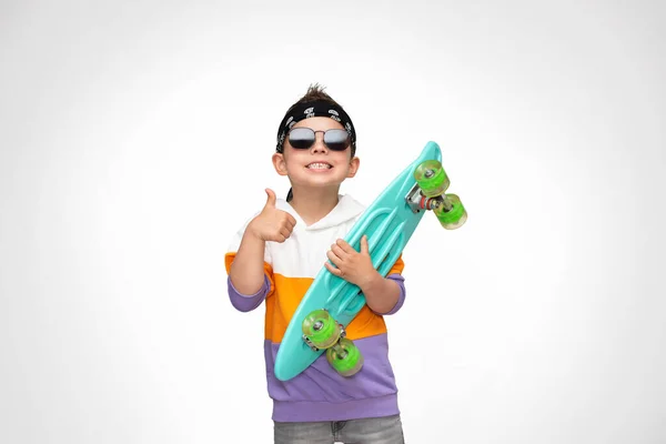 スタイリッシュな服とサングラスを身に着けている小さなクールなスケーターの男の子は彼の手の中にターコイズスケートを保持し 親指を示しています — ストック写真