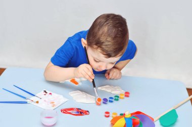 Mavi tişörtlü küçük bir çocuk akrilik boyalı alçı figürler çiziyor..