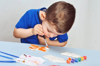 Mavi tişörtlü küçük bir çocuk akrilik boyalı alçı figürler çiziyor..