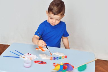 Mavi tişörtlü küçük bir çocuk akrilik boyalı alçı figürler çiziyor..