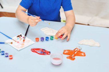 Mavi tişörtlü küçük bir çocuk akrilik boyalı alçı figürler çiziyor..