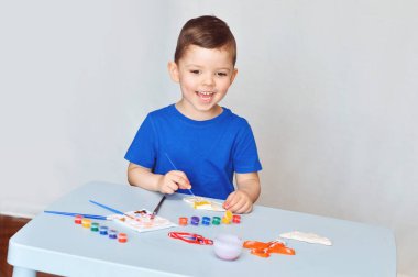 Mavi tişörtlü küçük bir çocuk akrilik boyalı alçı figürler çiziyor..