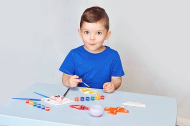 Mavi tişörtlü küçük bir çocuk akrilik boyalı alçı figürler çiziyor..