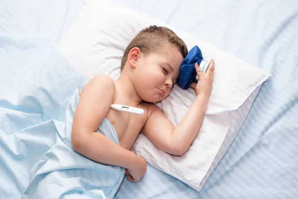 頭痛のある病気の小さな子供の男の子は彼のベッドの中にあり 温度を測定し 冷たいパックで頭を冷ます — ストック写真