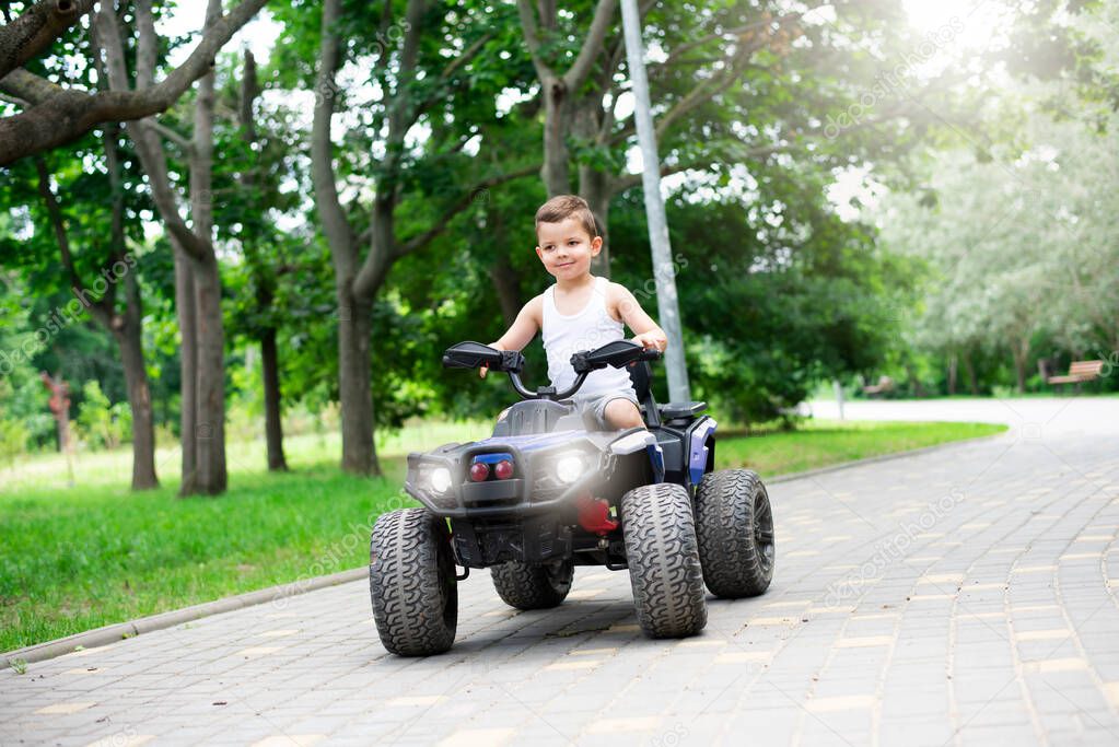 A cute five year old boy rides a black and purple ATV Quad bike in a summer park.