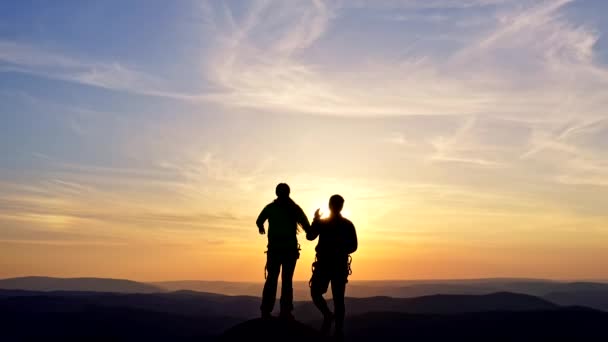 一对年轻夫妇在日落时站在山顶上高兴地举手. — 图库视频影像