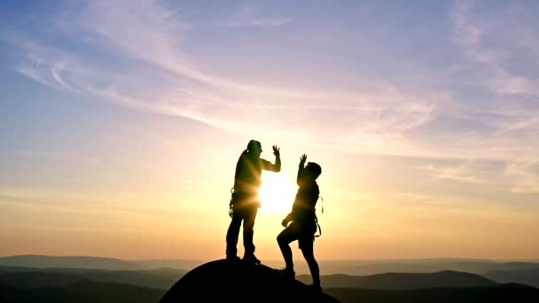 Silhouetten van een man en een vrouw geven hoge vijven op de top van een rotsachtige bergketen bij zonsondergang. — Stockvideo