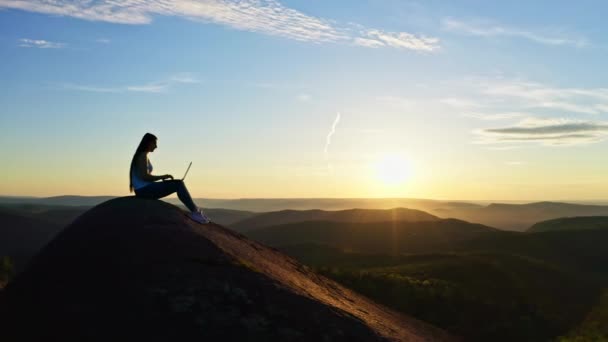 Scatti aerei di una silhouette di una giovane donna freelance che lavora su un computer portatile seduta su una montagna rocciosa al tramonto. — Video Stock