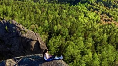 Kayalık bir çıkıntının kenarında otururken dizüstü bilgisayarda yazan genç ve çekici bir kadının hava görüntüsü.