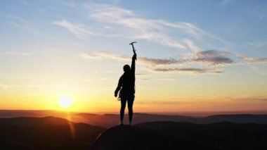 Yavaş çekimde, bir dağın tepesinde duran, kollarını kaldıran ve elinde bir buz baltası tutan ve gün batımında eğlenen genç ve mutlu bir kadın tırmanıcının görüntüsü..