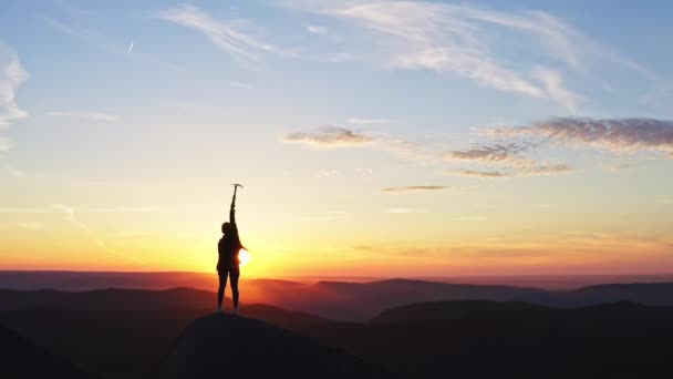 Slow motion silhouette of a female climber joyfully lifts an ice axe up celebrating victory at sunset. — Stock Video