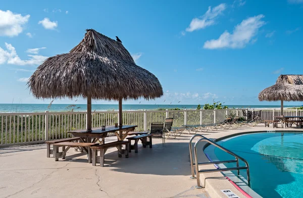 Beach table with hay parasol next to a swimming pool — Stock Photo, Image