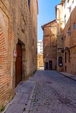 A narrow Segovia street is lined with historic buildings and wrought-iron balconies. The sunlit alley features architectural details and a lone figure, all beneath a bright blue sky.