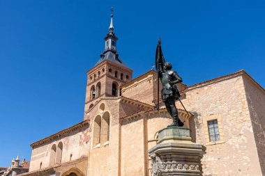 The statue of Juan Bravo stands before the Romanesque Church of San Martn in Segovia. The historic landmark, featuring a tall tower, is beautifully framed against a clear blue sky.