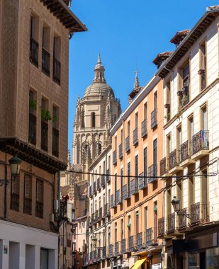 A narrow Segovia street is lined with historic buildings and wrought-iron balconies. The sunlit alley features architectural details and a lone figure, all beneath a bright blue sky.
