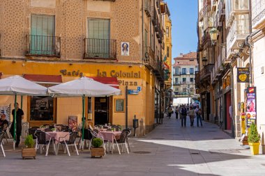 Downtown Segovia is bustling with life and activity. The pedestrian street features outdoor restaurants and caf tables set beneath a bright blue sky, surrounded by historic, patterned facades.