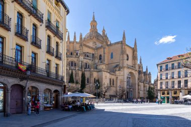 Segovia's massive Cathedral of Santa Mara dominates the plaza. Its Gothic style towers over the bustling public space, where Segovian life unfolds under a blue sky