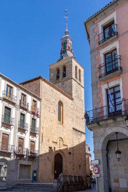 The Church of San Miguel's historic steeple rises above old Segovian buildings. The Romanesque tower dominates the bustling street scene under a clear blue sky